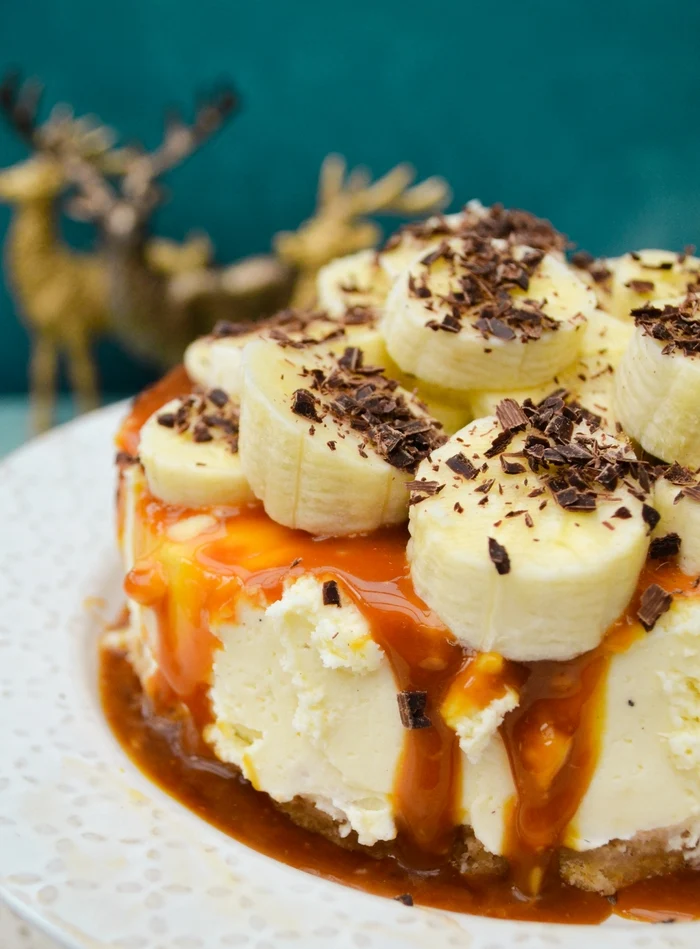 Close up Photo of a luxurious vegan banoffee pie on a cake stand with caramel oozing down the side, topped with slices of banana and grated chocolate.