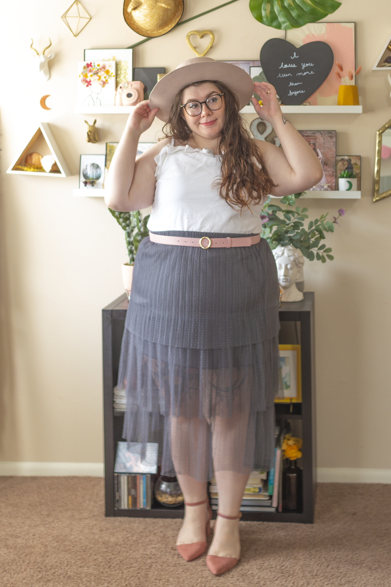 An outfit consisting of a pale pink wide brim pork pie hat, a white denim tank top with ruffled neckline tucked into a light grey pleated tiered midi skirt belted pastel pink belt and pastel pink pointed toe ankle strap heels.