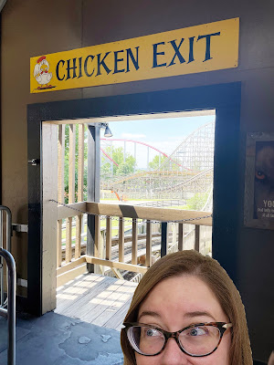 A white woman in glasses peeks in from the bottome with only her eyes and nose showing. She stands in a doorway with a rollercoaster in the background. Above the doorway is a sign that says "Chicken Exit."