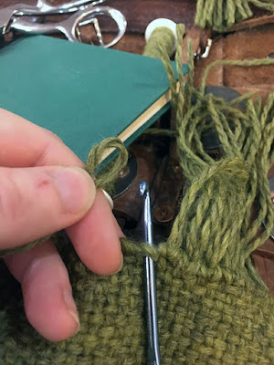 In the foreground, a white hand pinching a loop of light green yarn just left of a steel blue crochet hook inserted through the edge of a light green piece of cloth. The cloth is fringed at right. In the background, a slim, unmarked blue-green book, a white plastic bobbin of light green yarn, and the handles of silver sewing shears are piled on rust-brown woven leather armour.
