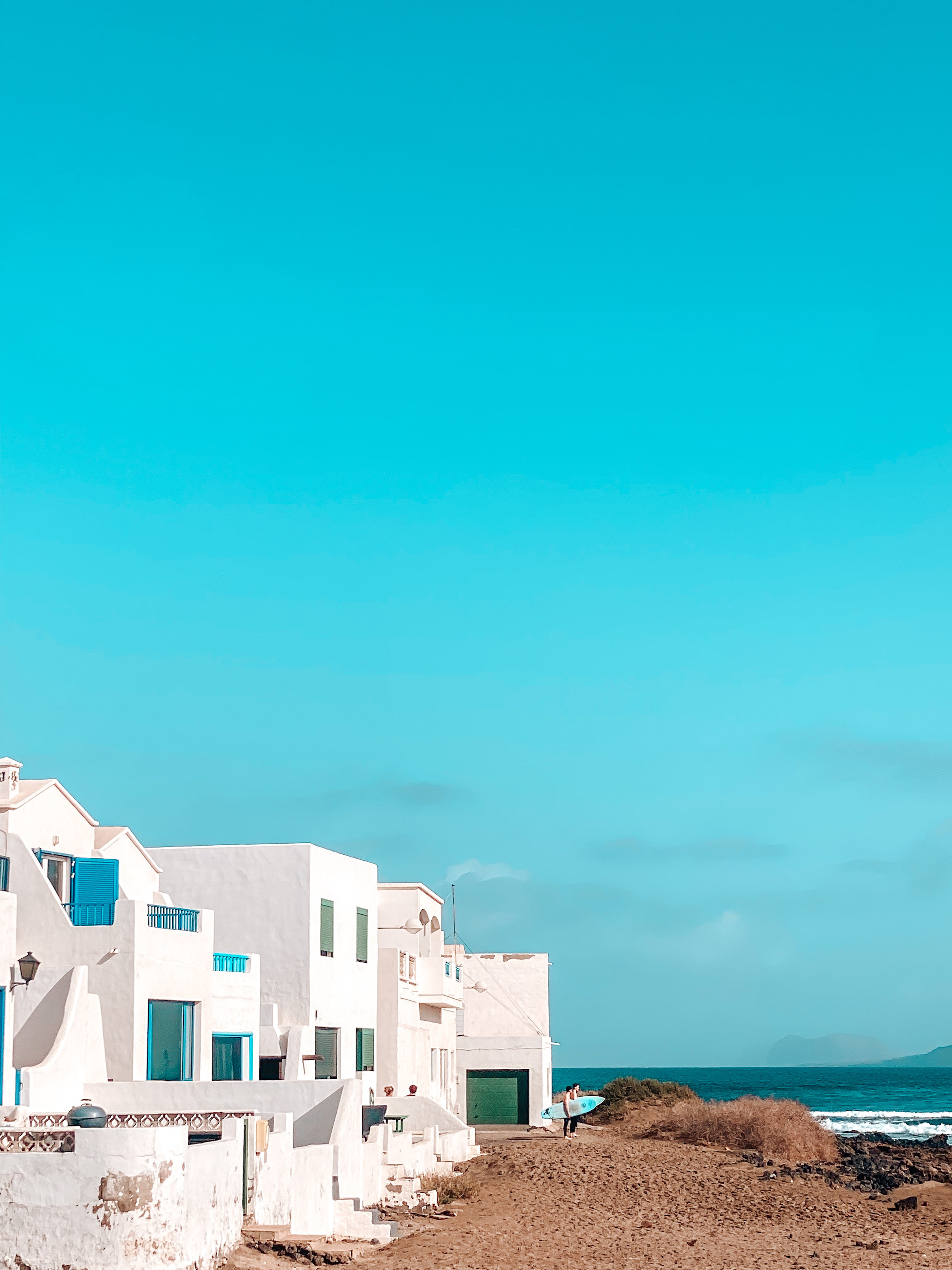 maisons blanches avec des volets bleus sur la plage de famara et surfers avec leur planche à la main en arrière plan