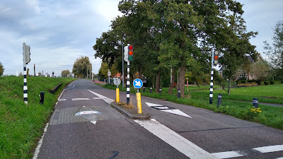 Traffic signals which resemble a pedestrian crossing with a skinny island between each traffic direction. There are detector loops showing in the surfacing and a triangular metal feature set flush to the road on each approach.