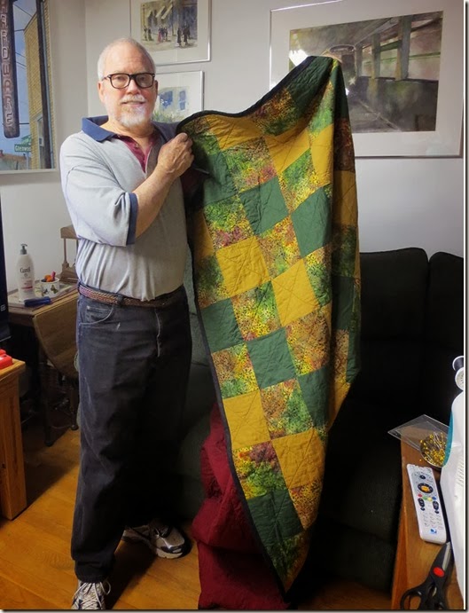 James holding quilt. Quilt comprises 6-inch squares of green, amber, and batik fabric, with maroon backing.