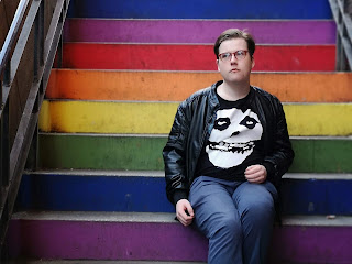 A close up of a guy staring outwards with round brown and green glasses with quiffed light brown hair, black leather jacket, blue shirt, black tshirt with a white skull on the front of it and bright blue chinos sat on rainbow coloured steps on a brigth background.