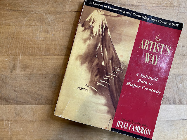 Picture of the book The Artist's Way, red cover with white and gold lettering, by Julia Cameron on a light wooden table