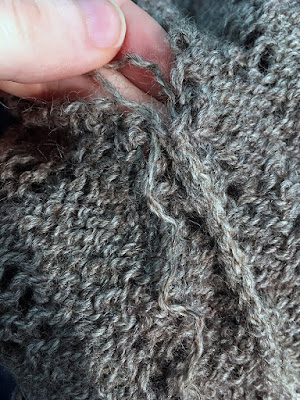 A close-up of a raised seam on the backside of grey stockinette lace fabric, with a tail partially pulled free of the pair of stitches at the open top of the seam, held between finger and thumb of a white hand.