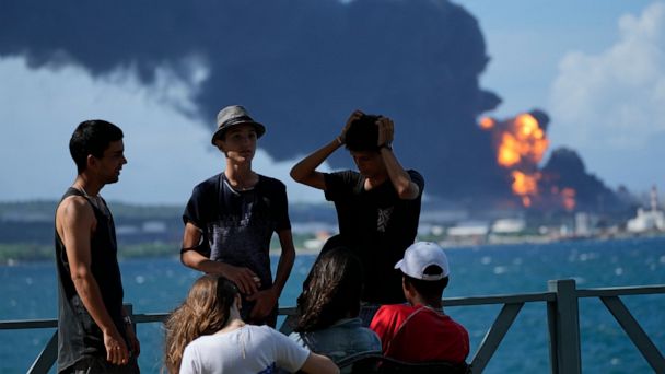 Youth assemble on a dock while flares and smoke keep on ascending from the Matanzas Supertanker Base