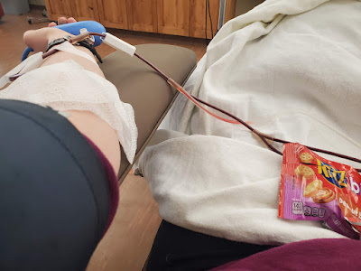 My arm hooked up to donate platelets. The blood pressure cuff is visible and there is a blanket and snack on my lap.