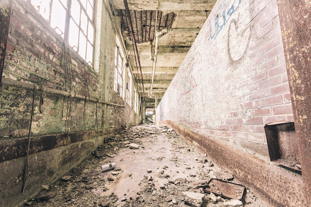 An abandoned bakery corridor with crumbling brick walls and decaying pipes. Light streams in through tall, grimy windows, casting shadows on the debris-strewn floor. Peeling paint and graffiti hint at the passage of time and the building's forgotten stories.