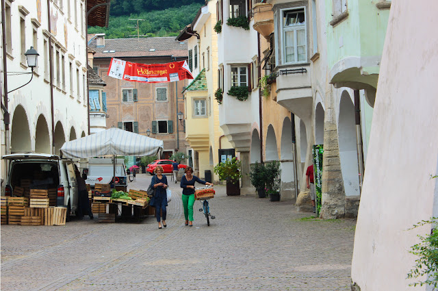 Przy Via Portici w Neumarkt-Egna wznoszą się wysokie kamienice z podcieniami. Fasady niektórych kamienic zdobią freski. Przy Via Portici kończy się właśnie targ, na którym sprzedaje się lokalne produkty. Pod białym parasolem stoi ostatni ze straganów, oferujących ułożone w drewnianych skrzynkach warzywa. Brukowaną ulicą idą dwie kobiety. Jedna z nich niesie torbę z zakupami. Druga prowadzi rower z wiklinowym koszem na kierownicy. Kosz dekorują czerwone kwiaty. 