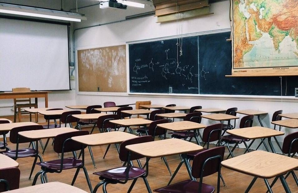 A imagem mostra uma sala de aula vazia com mesas, cadeiras e lousa