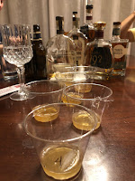 A kitchen table with a collection of bourbon bottles, wine glass, and plastic cups that are numbered for the bourbon tasting.