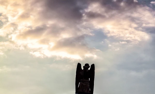 Un angel de cemente emergiendo hacia un cielo con nubes