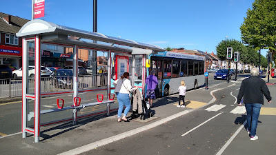 A floating bus stop with a glass shelter. People are getting on a white bus with a blue cab, including a woman with a buggy.