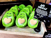 A photo of a large light borne square box containing some green aanthromorphised avocados smiling bubble bars next to a black rectangular card that says  Ava Cuddle Bubble Bar on a bright background