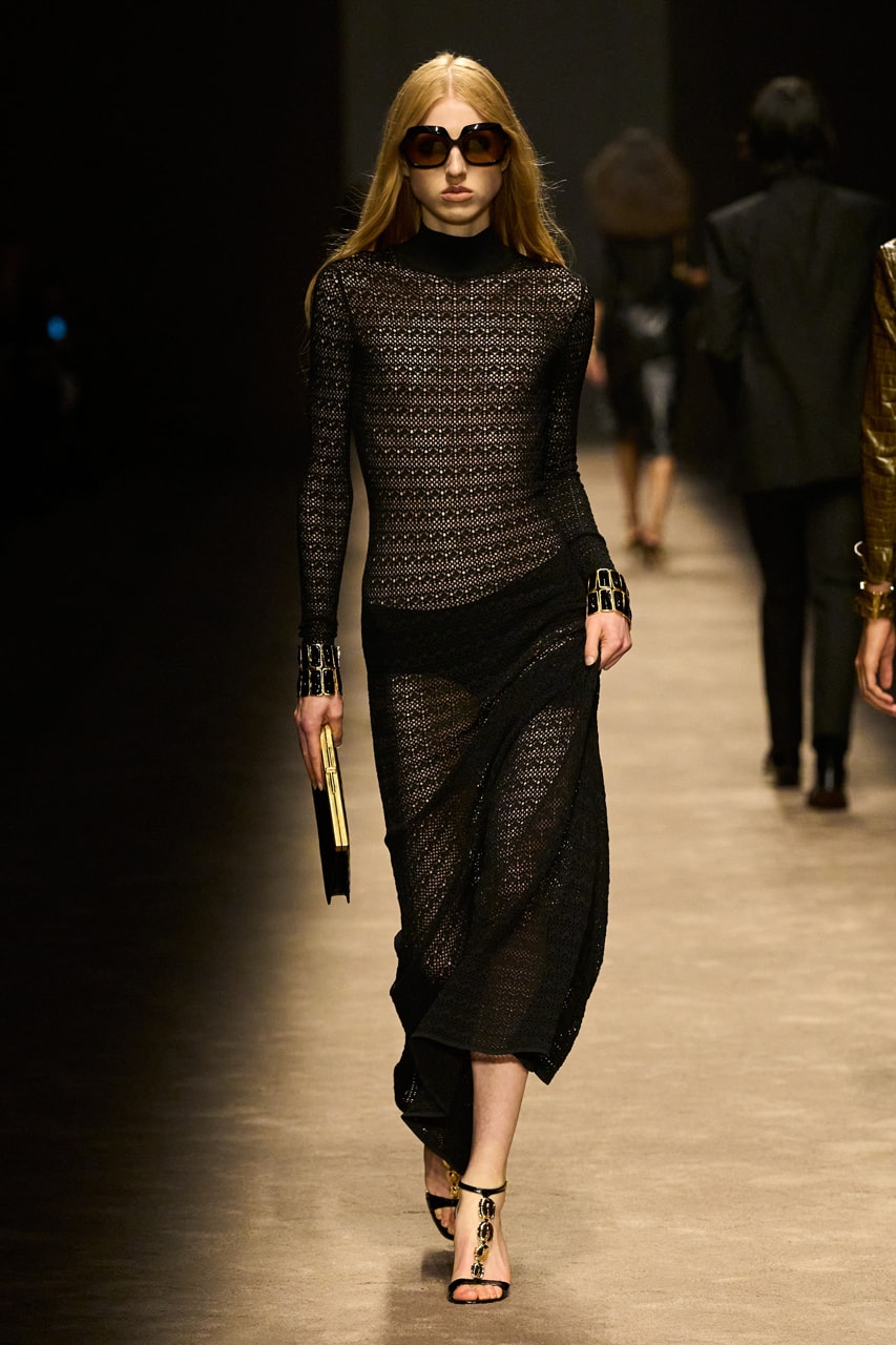 Woman with long gold blonde straight hair with a middle part wearing black oversized square sunglasses, black see-through crochet maxi dress, strappy black sandals with black stone details down the front, and chunky black stone bracelets on both wrists, carrying a thin black clutch with gold detailing, walking on a taupe carpeted runway