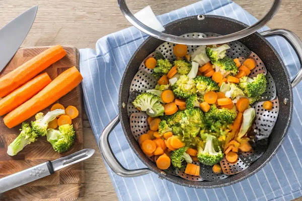 Freshly steamed broccoli and carrots in a steaming basket on a stovetop, showcasing a healthy cooking method to preserve nutrients. Includes whole carrots on a cutting board, emphasizing how to steam vegetables for maximum nutritional benefits and flavor.