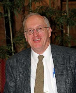 Smiling man with gray hair and glasses wearing a gray plaid blazer with white shirt and olive green tie