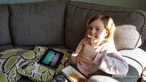 Emily, a little girl, is sitting on a couch eating waffles and syrup.  She is giving a big thumbs up.