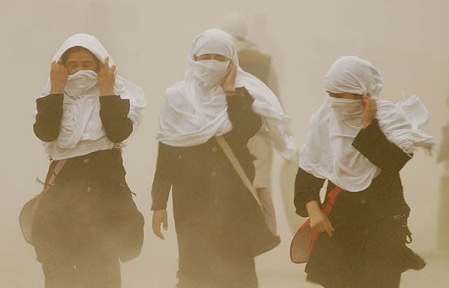 Fotografia de três mulheres utilizando roupas pretas e véu islâmico branco. Elas estão andando em direção à frente da imagem e  tentam segurar seus véus enquanto passam por uma tempestade de vento.