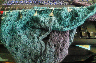 A gradient lace asymmetrical shawl knit up on a straight needle.  The long end is tucked under the end of the straight needle, and the gradient goes from purple to teal. 