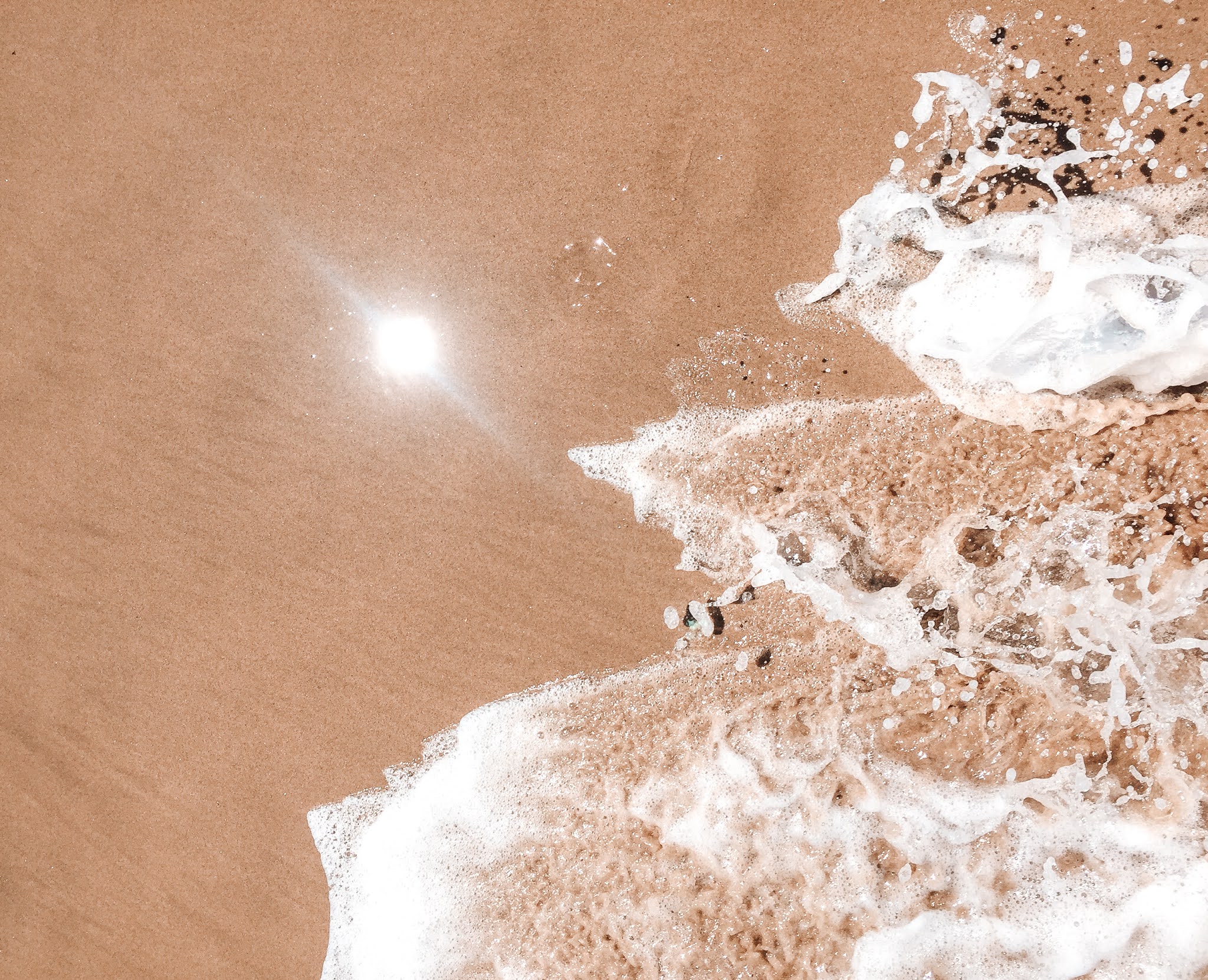sable brun avec le soleil qui se reflète sur le sol et l'écume amené par une vague en bas à droite