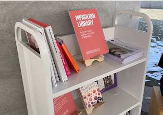 White metal book cart with books and a bright orange sign that says MPavilion Community Library