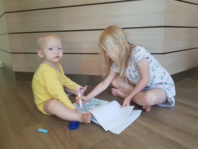 A toddler sitting on wooden floor next to her big sister while big sister colours in her colouring book