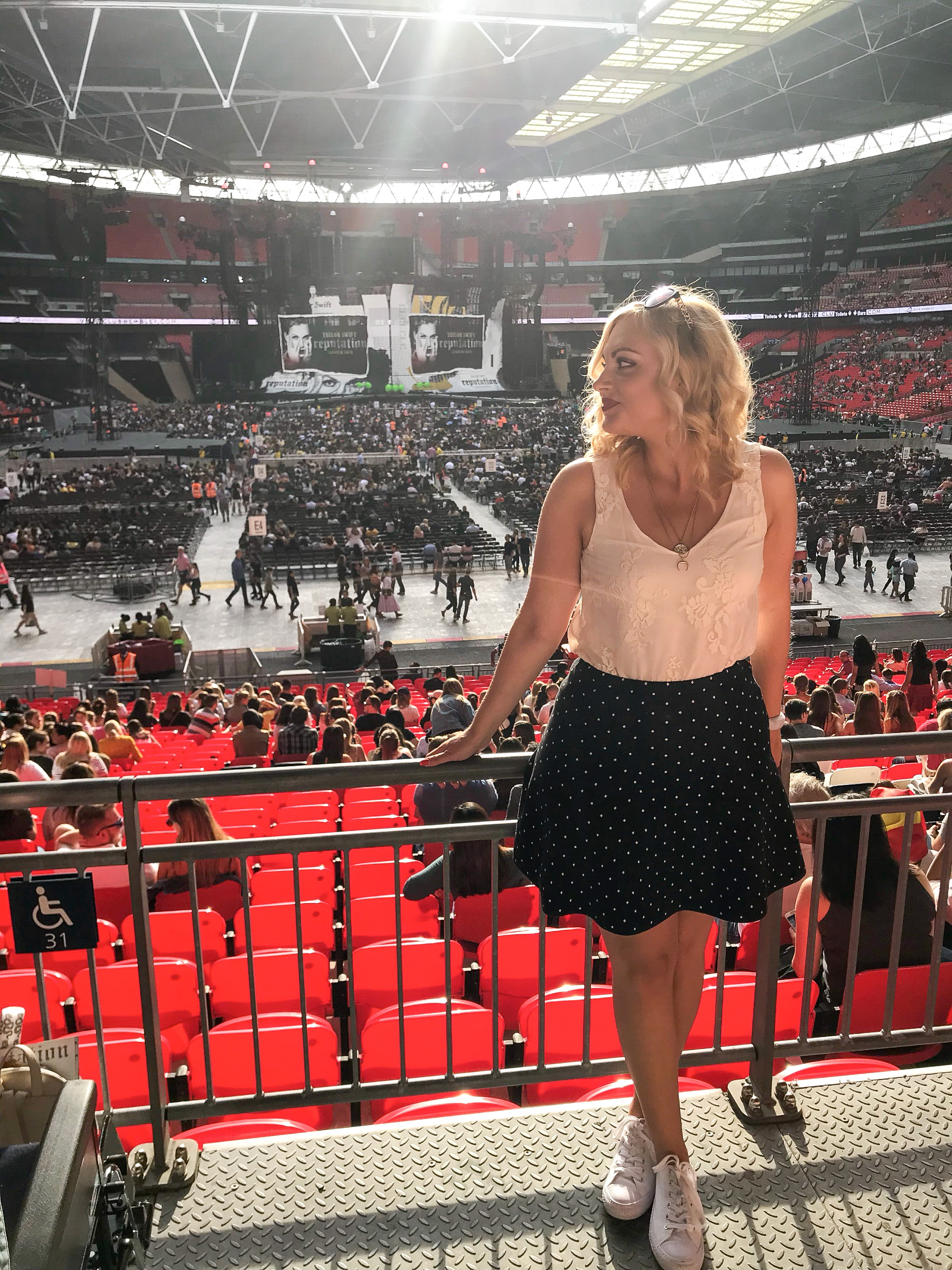 The Reputation Stadium Tour London 2018, Blonde girl stands in front of a barricade in Wembley stadium with Taylor Swift's Reputation Tour Stage behind her. She wears a cream lace camisole & black and white polka dot skirt.