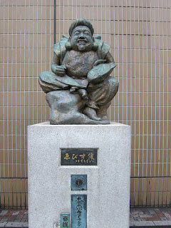 Ebisu, statue outside the Ebisu train station in Tokyo. A bronze statue of a small seated man, smiling and holding a fish, in front of a tiled wall.