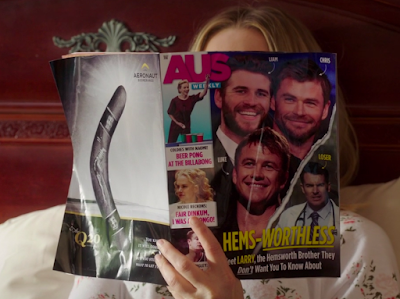 Eleanor holding an AUS Weekly magazine in front of her face. The main cover story is the one quoted above, with pictures of three Hemsworth brothers' faces, and down to the side, a picture of a man wearing a white coat with a stethoscope around his neck, with the word LOSER pointed towards him. The back of the magazine has an ad for a boomerang. Other cover articles: A picture of someone named Naomi playing beer pong, titled "COLDIES WITH NAOMI! BEER PONG AT THE BILLABONG"; a picture of Nicole Kidman's head, titled "NICOLE RECKONS: FAIR DINKUM, I WAS A DRONGO!" but Eleanor's finger is obscuring part of the text so the "A DRO" of "A DRONGO" was inferred by me