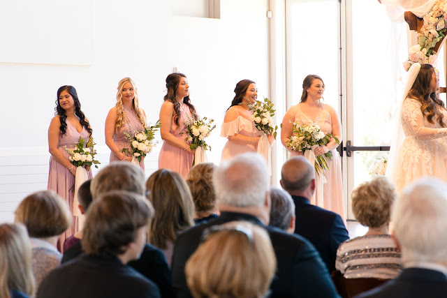 May 1st is a special day in the lives of Jonathan and Katelin, for it was on this day one year ago that he proposed at the Myriad Gardens in Oklahoma City.  Both Katelin and Jonathan grew up in Stillwater, went to the same middle and high school, but didn’t actually get to know each other until one of Katelin’s friends suggested that she invite Jonathan to the Senior Prom.  Katelin didn’t think that was a good idea at all, since she described him as “a very shy, nerdy kid,” and she was a “social butterfly.”  Little did she know that “we would really hit it off…and things have just gotten better every day since!”     And what was not to love about this day?  The bride and groom chose the Sparrow in Stillwater as their venue, with a rustic, architecturally modern barn, and 30 beautiful acres to roam in.   Katelin and her bridesmaids spent the morning, tucked away in their own getting-ready suite, finishing up with hairdresser, make-up artist, and assisting the bride into her gorgeous gown.  They were also trying to be a calming influence, but as the time for the First Look arrived, everyone was excited…and emotional.  Especially the newlyweds-to-be!     Katelin and Jonathan exchanged sweet letters during the First Look, which brought joyful tears to each of them, and then they were whisked off to create some magical couples’ portraits.  Wedding party pictures followed, as the bridesmaids gathered around Katelin, and the groomsmen’s personalities emerged as they enacted the “Mario” jump!    At last the hour came for the ceremony to begin.  The bridesmaids, dressed in pastel shades of pink and lavender, came down the staircase on the hand of their respective groomsman, while Jonathan waited patiently at the altar for his bride.  Added to the wedding party were the couple’s two Golden Retrievers—the “flower girl and ring bearer.”  The bride was stunning in her lace-embroidered, elegant wedding gown, carrying a lush floral bouquet consisting of white roses, white ranunculus, and lush greenery.     After the couple exchanged vows, guests were invited to participate in a buffet dinner while they enjoyed the festivities of the evening…the first dance, dances with the respective parents, toasts to the bride and groom, and dancing.  The culmination of the evening took place when Jonathan, in light-hearted fun, smashed some of the wedding cake on Katelin’s face, only to be sprayed with champagne when the bottle popped off as he was opening it!  True to her humorous good nature, Katelin took it all in stride!     This is one of those weddings we will remember, reminding us that soul mates do find one another in the most wonderful of unexpected ways…and we wish this delightful couple a lifetime of happiness together!