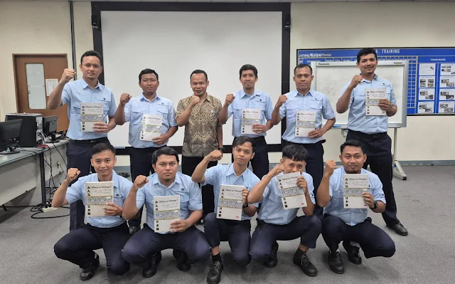 Sekelompok pekerja pria berpose sambil memegang buku pelatihan “Minna no Nihongo” di ruang training, mengenakan seragam biru muda.