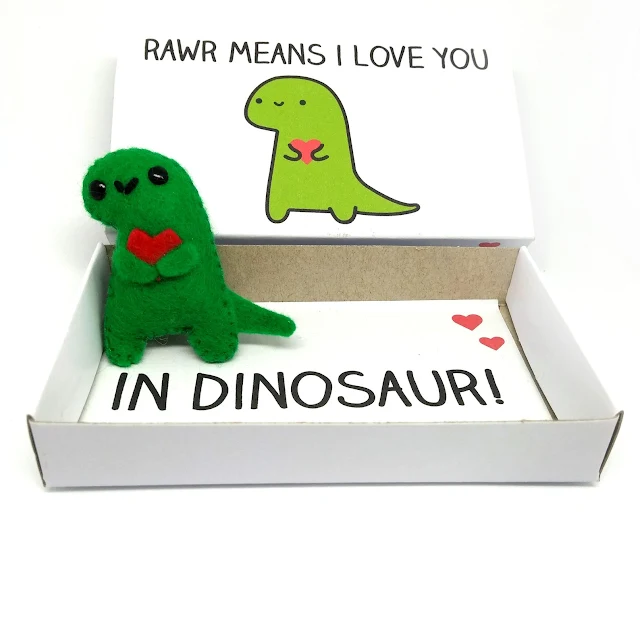 An image from the Etsy store selling a cute mini felt dinosaur magnet holding a heart which comes in a matchbox with the text Rawr means I love you in dinosaur