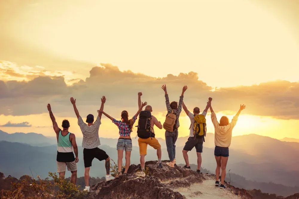 Group of happy friends stands with raised arms at sunset mountain and enjoys sunset