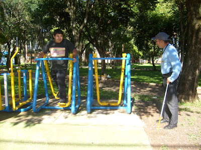 Foto em plano geral mostrando Marcos em pé em um dos aparelhos de ginástica, no lado direito da imagem está Luiz olhando em direção a Marcos. Ao fundo várias árvores.