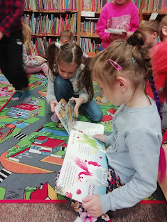 Na pierwszym planie dziewczynka czyta książkę. W tle dzieci i regał z książkami.