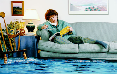 Photo of a woman talking on the phone and reading the phone book on her couch while the room she is in is flooded