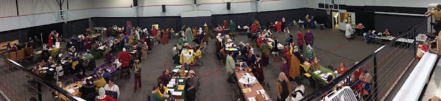 A panorama of a grey-carpeted gym full of long tables arranged in rows end-on to the viewer, with people in bright medieval clothing sitting and standing at the tables, talking to each other. Various items are on the tables, all accompanied by white sheets of paper, and purple pennants on short stands.