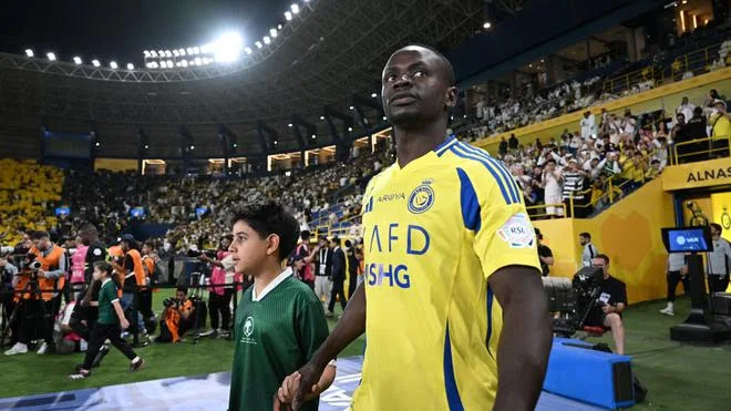 Imagem de um jogador de futebol famoso entrando em campo com uma criança durante um evento esportivo, torcedores no fundo. O jogador veste a camisa amarela do Al Nassr.