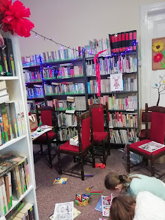Z lewej stony fragment regału z książkami. Na drugim planie widać siedzące na podłodze dziewczyny. Kolorują. W lte regały biblioteczne.