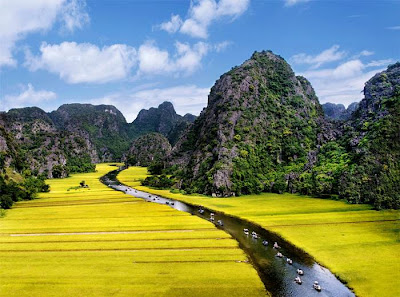 The Tam Coc (“three caves”) portion is a three-hour excursion by small boat along the Ngô Đồng river,  beginning at the village of Van Lam and proceeding through a scenic landscape dominated by rice fields and karst towers. The route includes floating through three natural caves (Hang Ca, Hang Hai, and Hang Ba), the largest of which is 125m long with its ceiling about 2m high above the water. The boats are typically rowed by one or two local women who also sell embroidered goods.