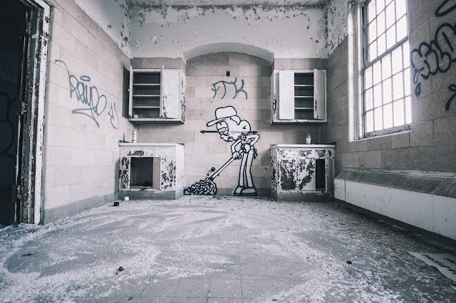 An abandoned room with flaking walls and graffiti, including a caricature of a figure sweeping the floor.