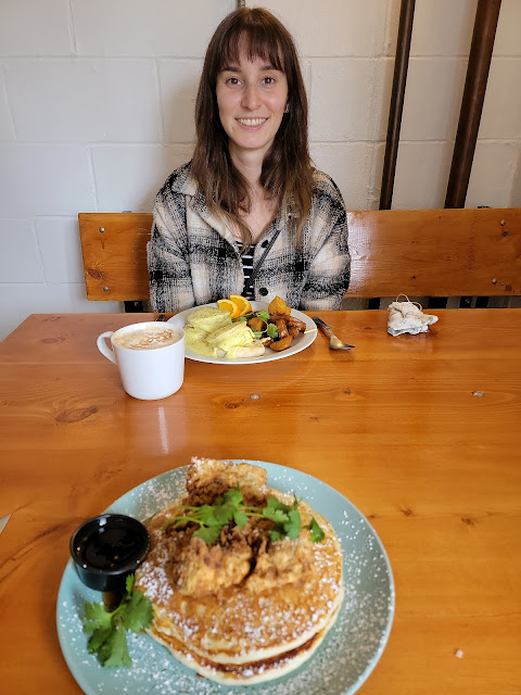 Me sitting in front of vegan eggs benny and a pumpkin spice latte. Across from me is a plate of vegan fried chicken and pancakes.