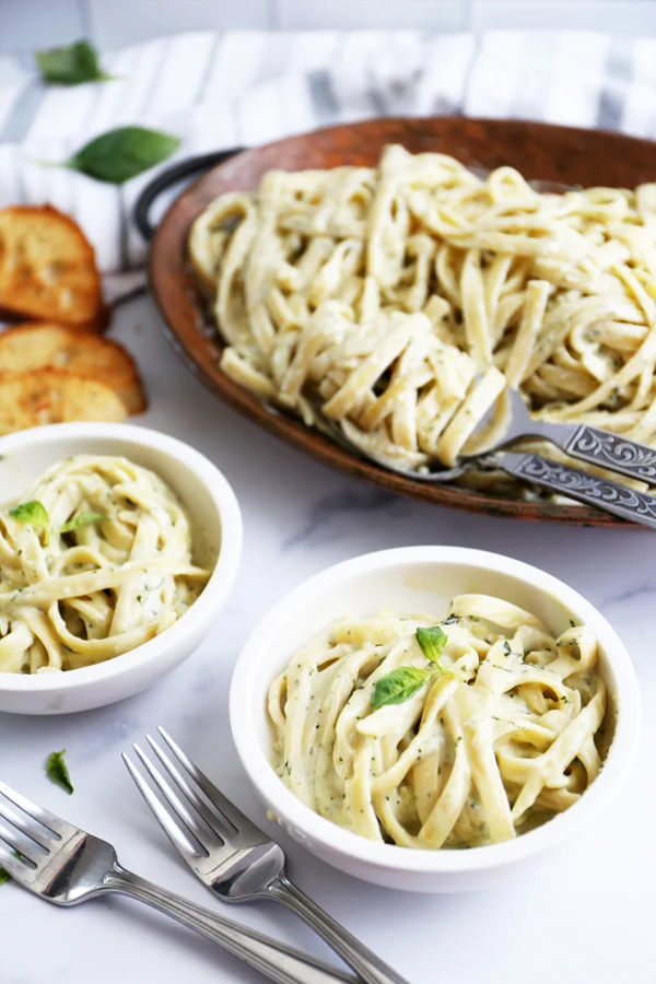 A rustic serving tray showcasing creamy pesto pasta, served in two bowls. The pasta is garnished with fresh basil leaves, creating a visually appealing presentation. A side of garlic bread complements the dish.