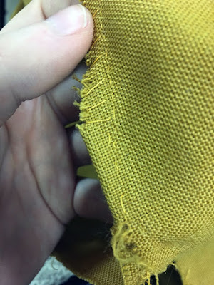 A close-up of a tiny folded section in mustard-brown fabric between two nearly-intersecting seams, stitched with bright-yellow thread and fniished with huge whipstitches, held up by a white hand.