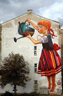 Natalia Rak e seu gigantesco grafite em Bialystok, Polônia.A foto mostra um gigantesco grafite na parede lateral de um prédio. O grafite à direita é de uma garota ruiva com trancinhas e traje típico polonês: Vestido regata com corpete preto, saia listrada em tons de vermelho, azul, amarelo e branco. O vestido tem um grande laço vermelho na cintura e está sobre uma camisa branca de mangas curtas bufantes com bordados. A garota parece flutuar, está descalça, na ponta dos pés regando uma árvore natural à esquerda, junto à parede do prédio.