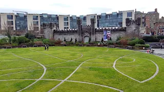Large lawn of bright green grass with a design of many curves made out of small grey stones. Beyond the grass is a piece of a castle wall. Behind the castle wall are modern looking short buildings made of glass and concrete