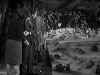 A man and a woman stand together, looking off the edge of a cliff at a crashed spaceship, broken in two in a barren area.