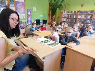 Pani bibliotekarka pokazuje dzieciom książkę o wiośnie. Dzieci siedzą naprzeciwko pani przy stolikach Tło: regały z książkami.